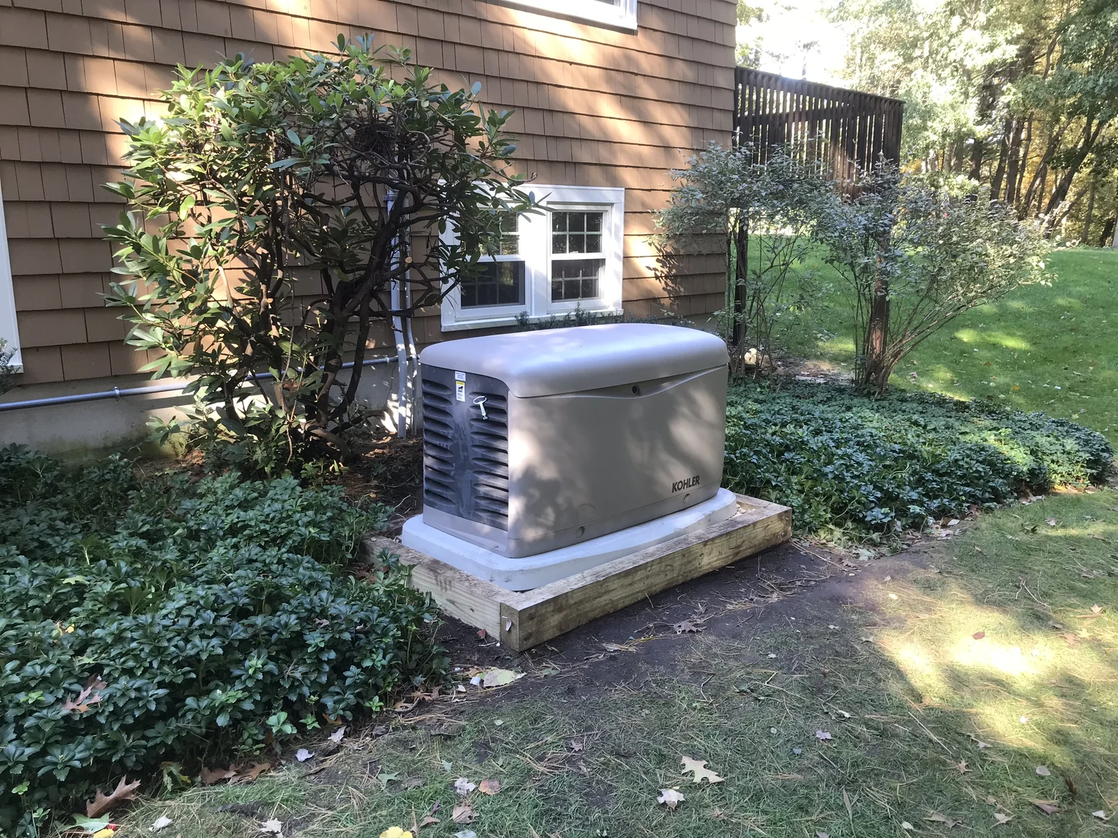 A standby generator on a pad beside a shingled house and green planting.